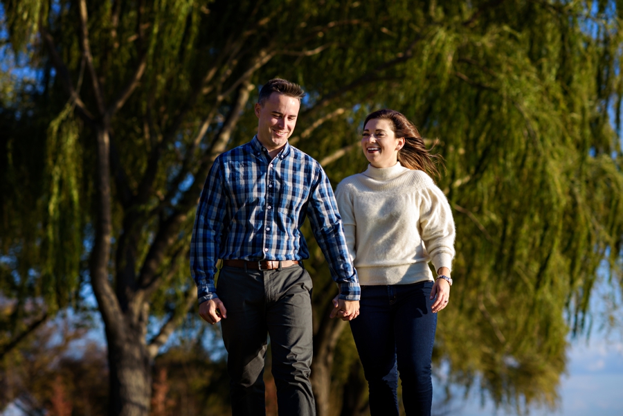 An engaged couple walk along the tidal basin with weeping willows blowing in the wind behind them.